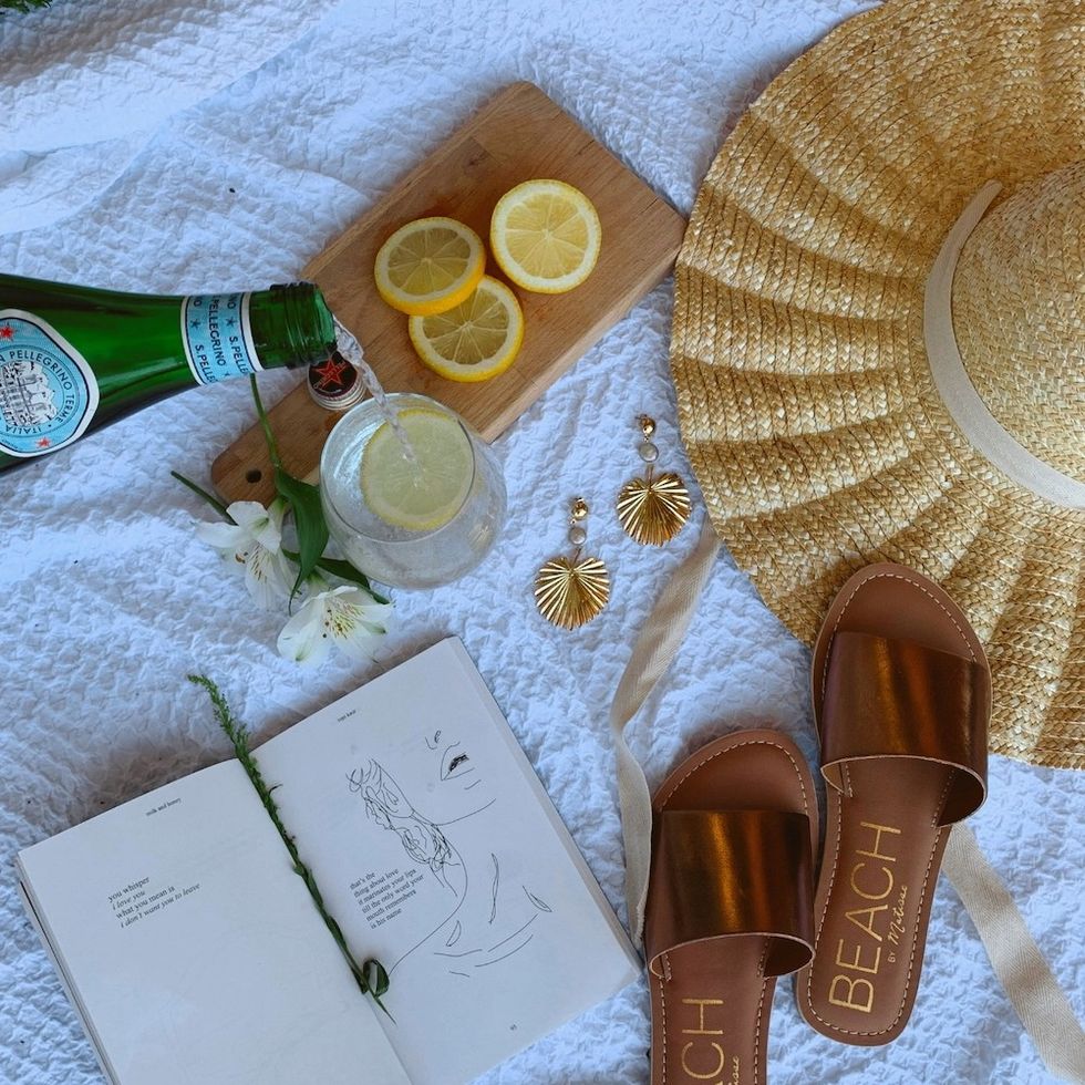 a beach blanket with a book, shoes, a hat, and a drink on it