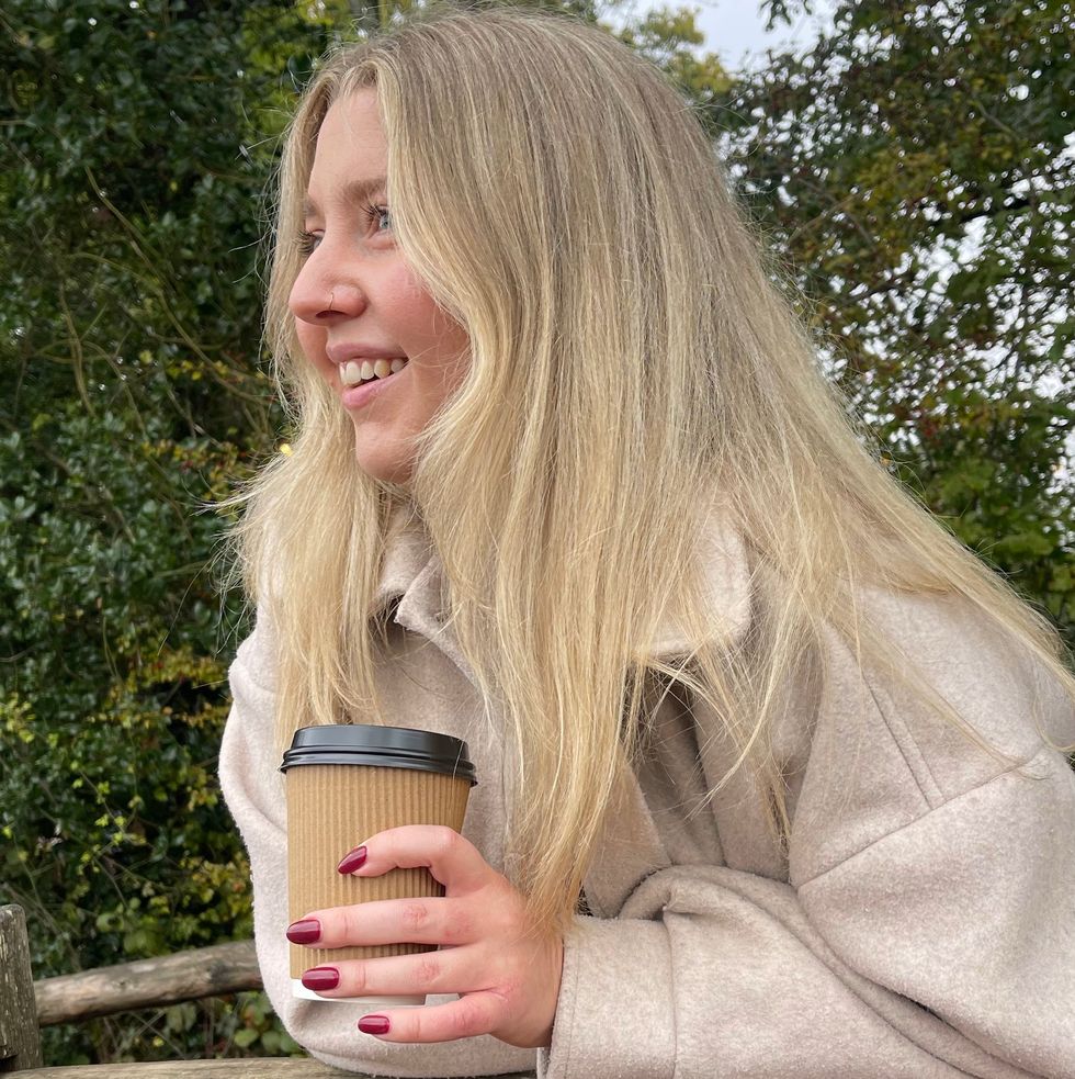 a blonde woman in a fall coat holding a coffee in the park