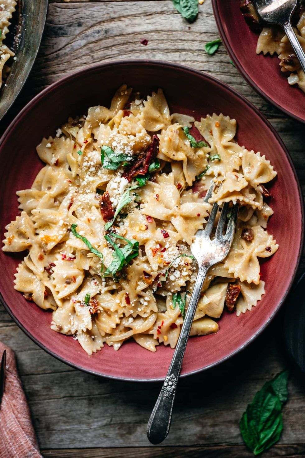 A bowl of sun-dried tomato pasta sits inside of a red bowl with a fork.