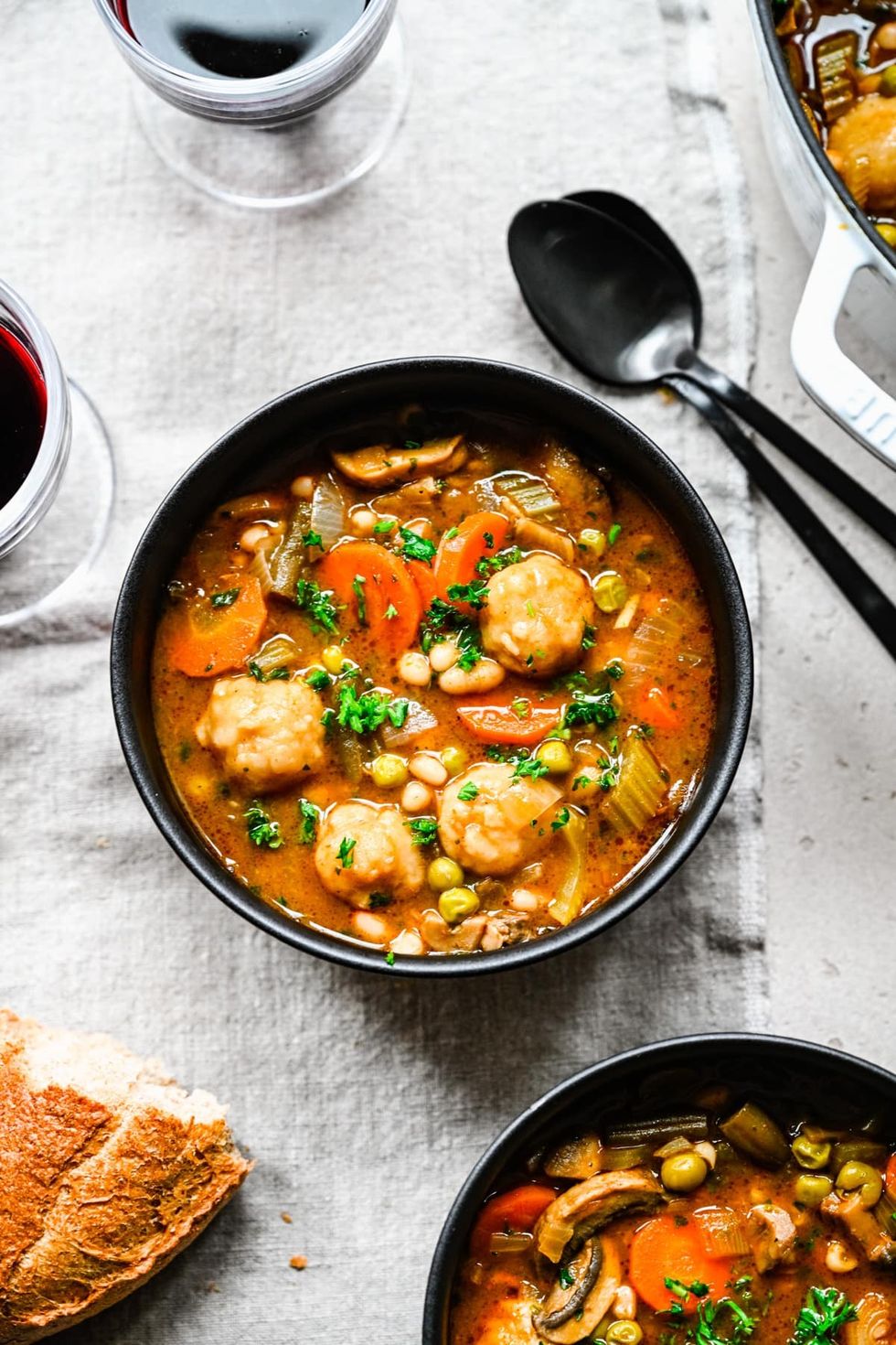 A bowl of vegan stew and dumplings sits next to two spoons and a piece of bread.