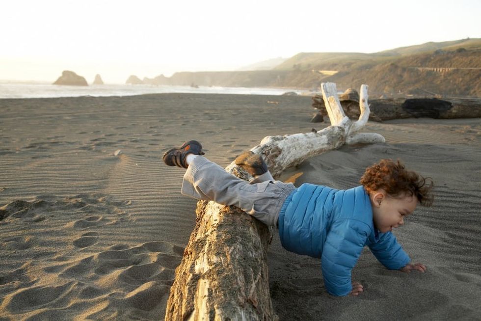 A boy crawls over a log on a beach