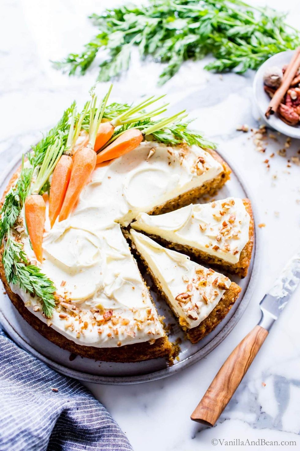 A carrot cake, garnished with full carrots, sits on a cake plate with a serving knife next to it.
