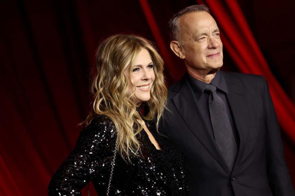 A couple in formal attire smiles on a red carpet with a red draped background.