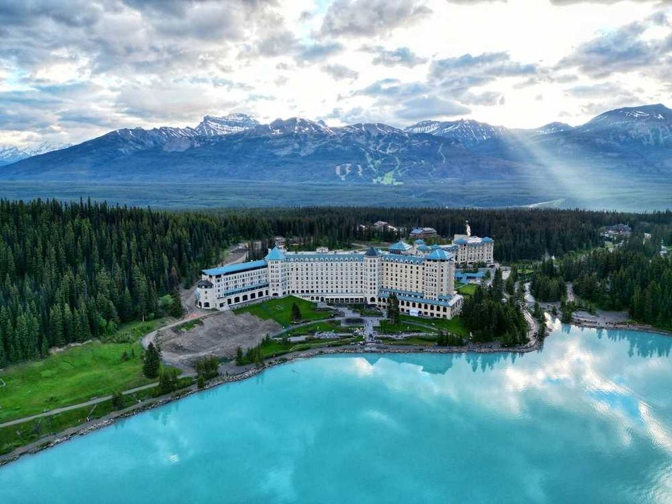 A grand hotel by a turquoise lake, surrounded by forest and mountains under a cloudy sky.