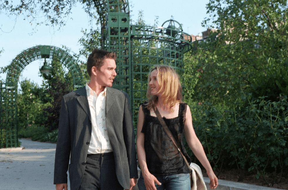 A man and woman walking and talking in a lush, leafy garden with green archways.