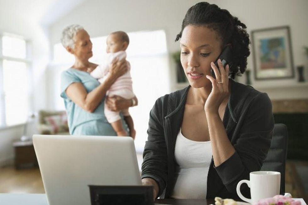 A mother works on her phone and computer while her nanny holds her baby in the background