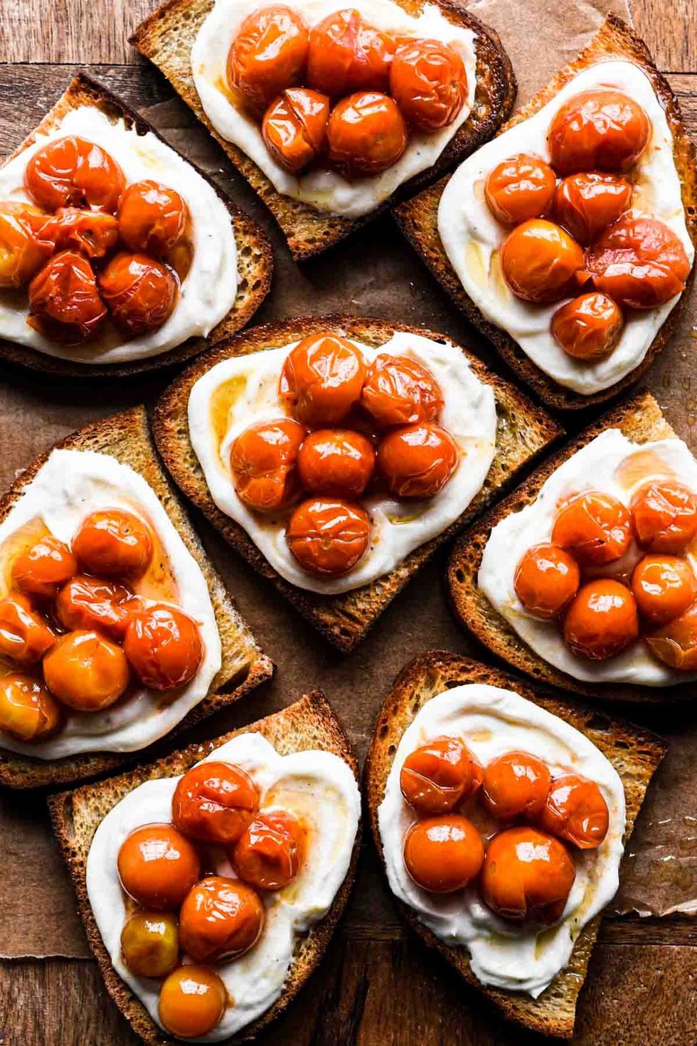 A number of tomato confit and whipped ricotta toasts are on a brown surface.