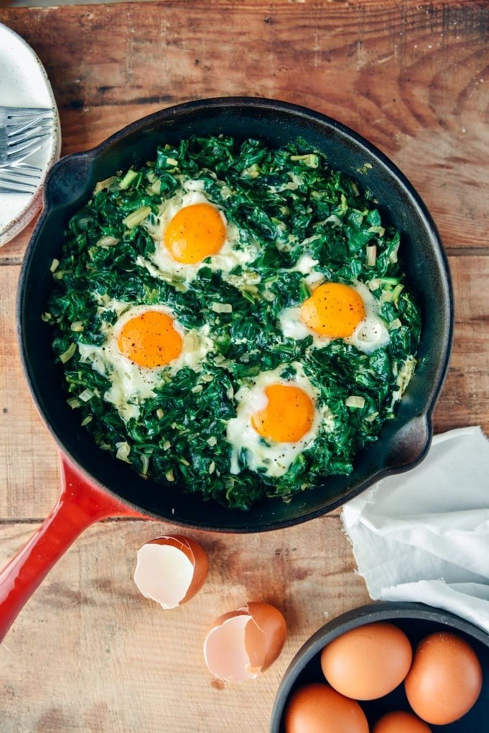 A pan of fried eggs with spinach sits on a wooden surface next to a bowl of eggs.