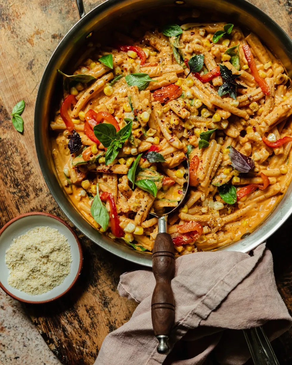 A pot of vegan red pepper pasta with charred corn and basil sits on a wooden surface.