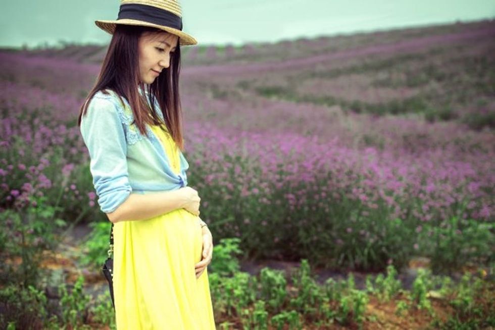 A pregnant woman in a bright yellow dress stands in a field of purple flowers