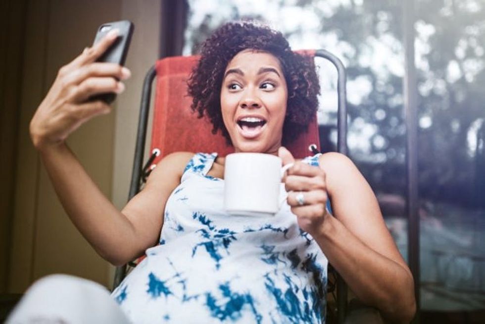 A pregnant woman smiles as she holds a mug and takes a selfie