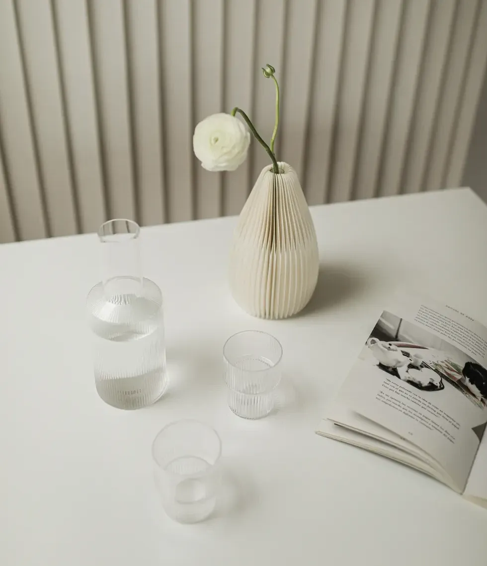 A white vase with a rose sits on a white table next to a water pitcher and two glasses.