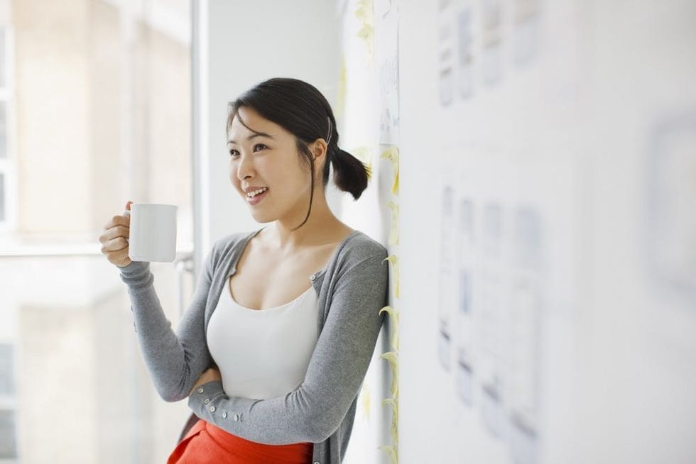 A woman drinks coffee while leaning against a whiteboard