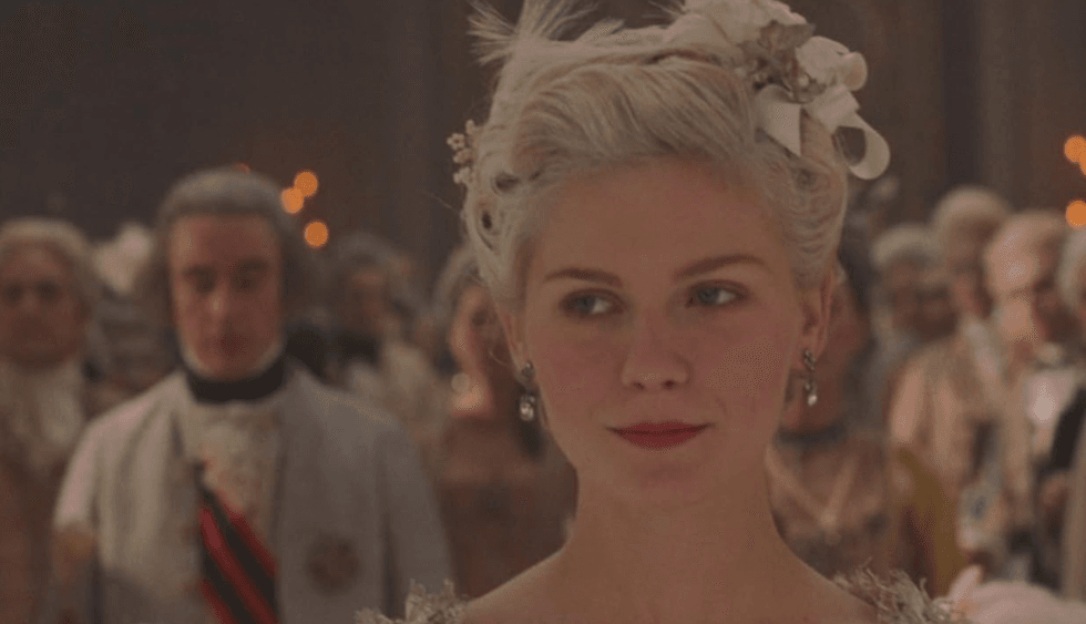 A woman in elegant historical attire at a formal gathering, with guests in the background.