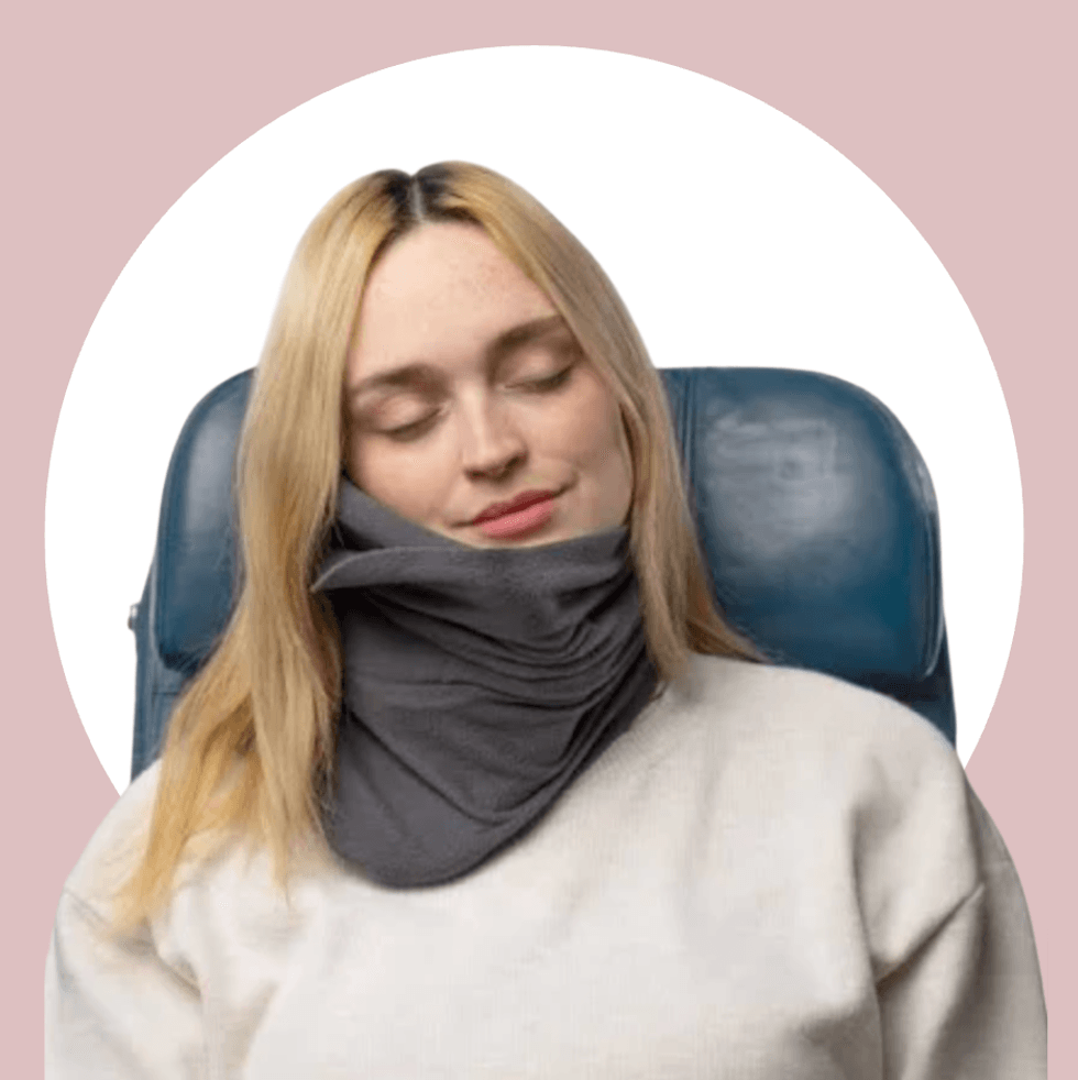 A woman rests with eyes closed in a blue chair, wearing a gray neck pillow and cream sweater.