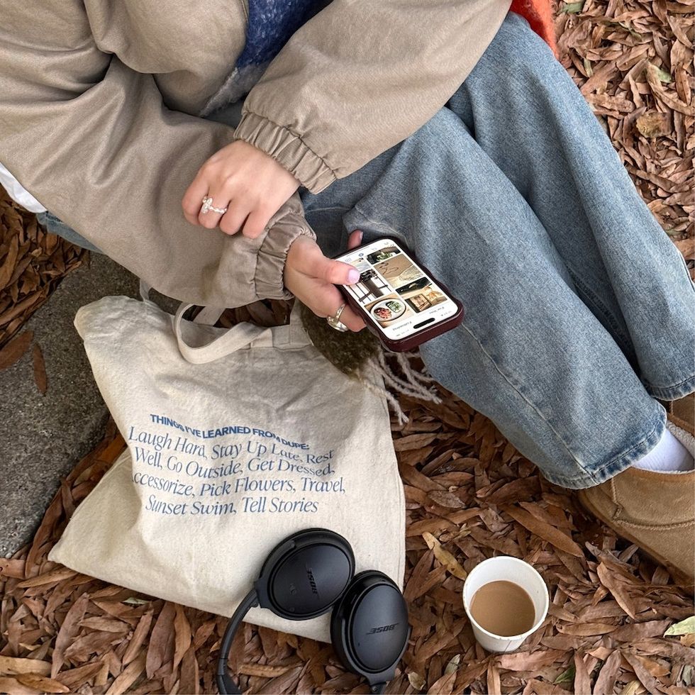 a woman sitting on the curb with the fall leaves, her bag, her headphones, a coffee, and looking at her phone