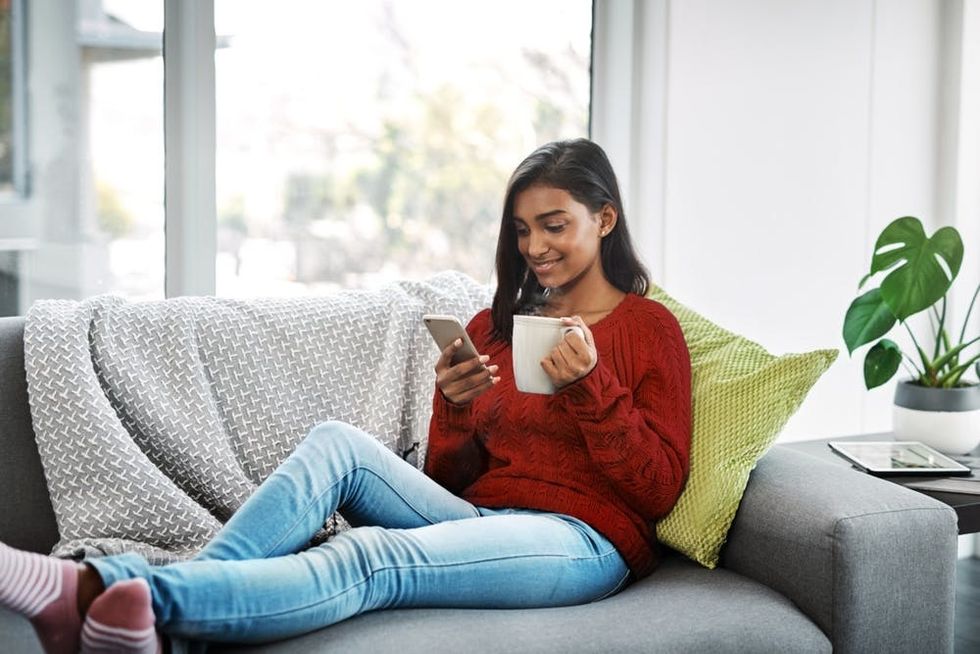 A woman smiles while using a phone and drinking a hot beverage
