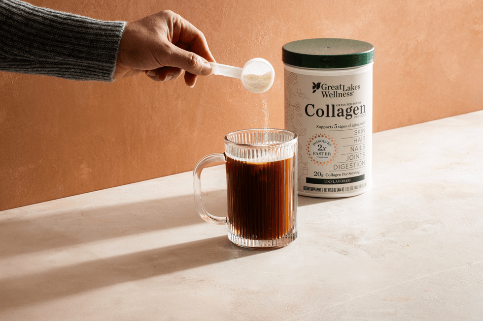 Adding collagen powder to a glass mug of dark liquid next to a collagen container.
