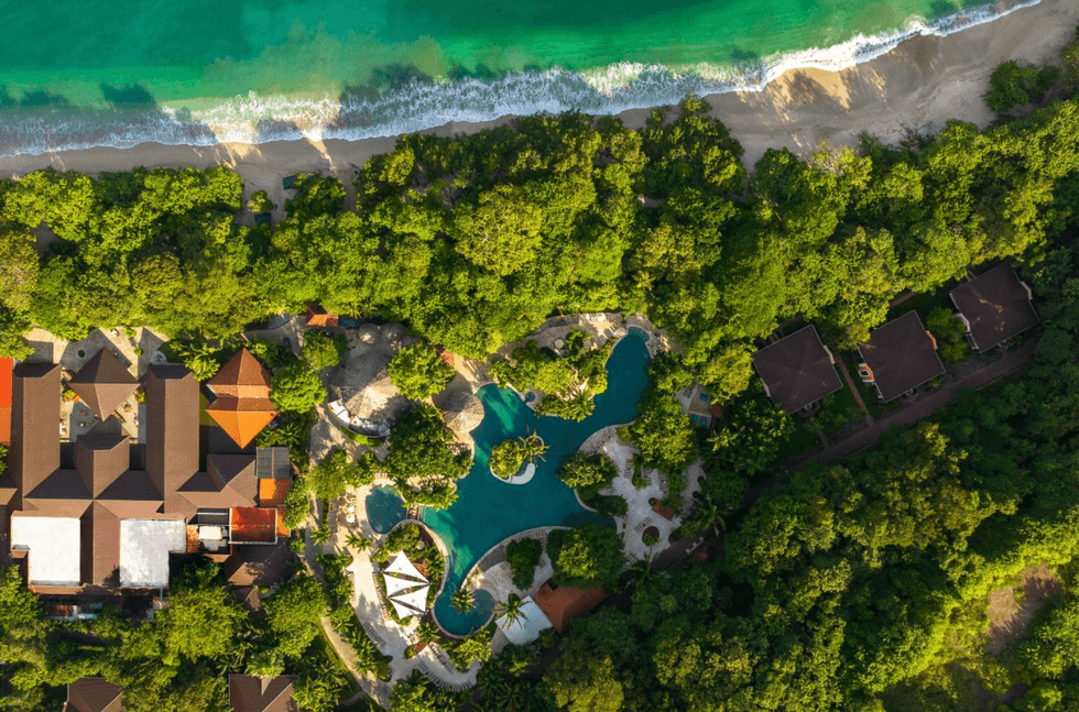 Aerial view of a coastal resort with lush greenery, a pool, and beachfront.