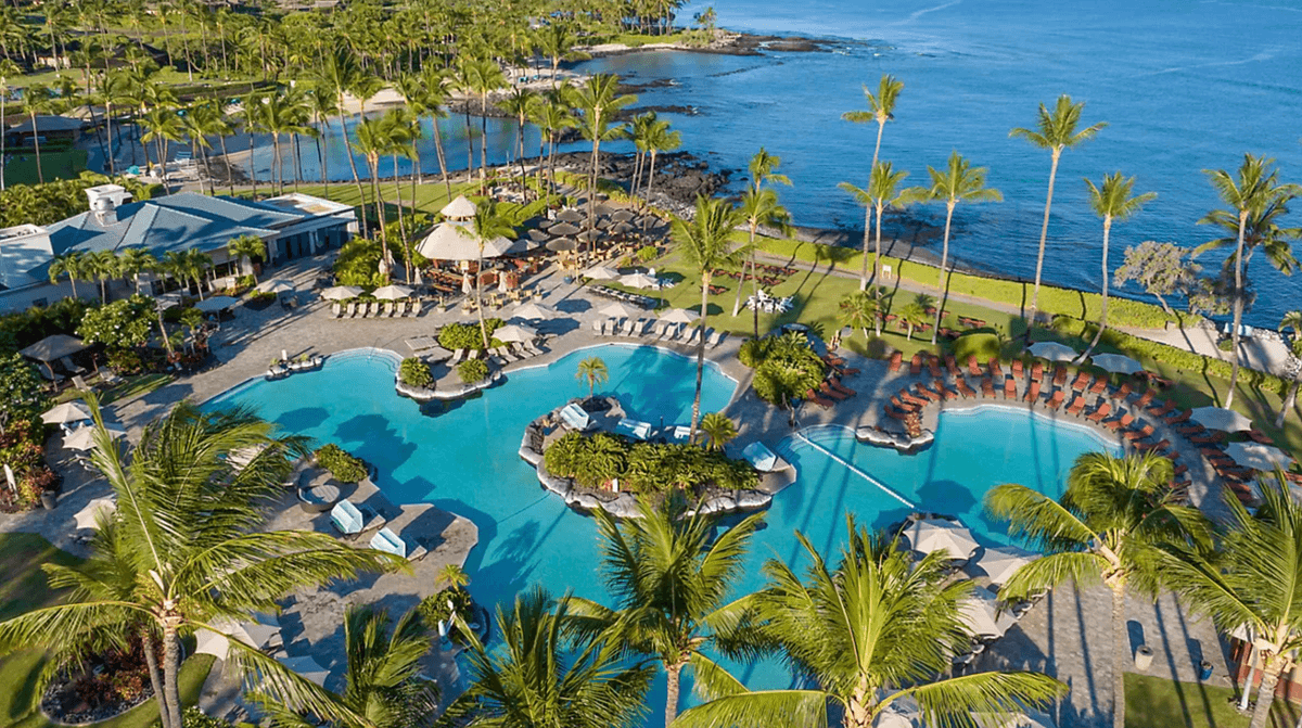 Aerial view of a tropical resort pool, palm trees, and ocean coastline.