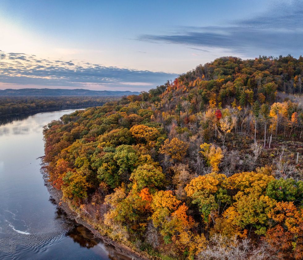 Aerial view of autumnal forest by a wide, calm river under a partly cloudy sky.