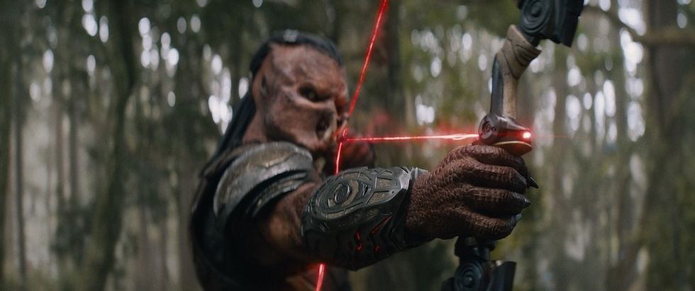 Alien archer aims high-tech bow with red lasers in a dense forest.