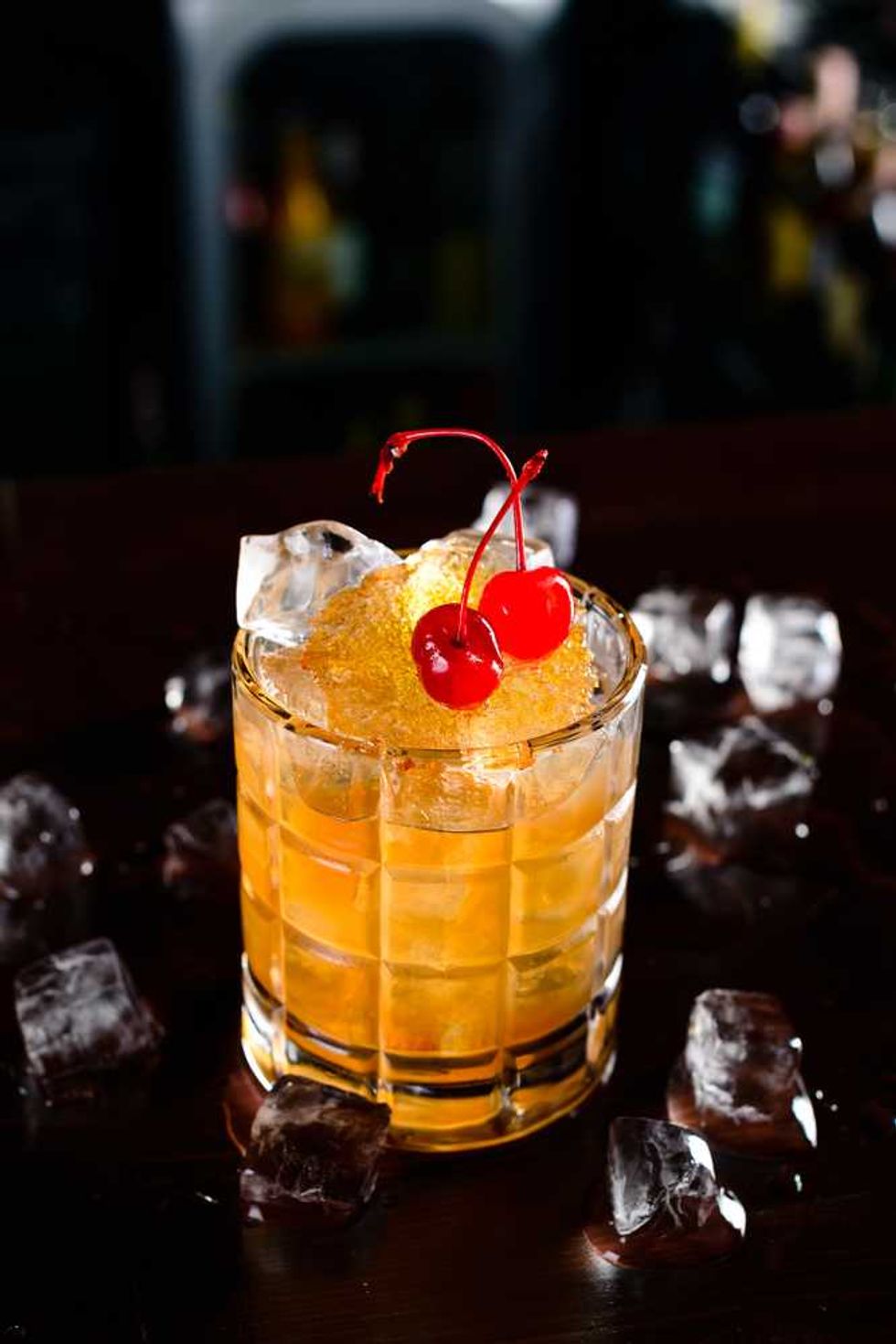 Amber cocktail in a glass with cherries and ice cubes on a dark surface.