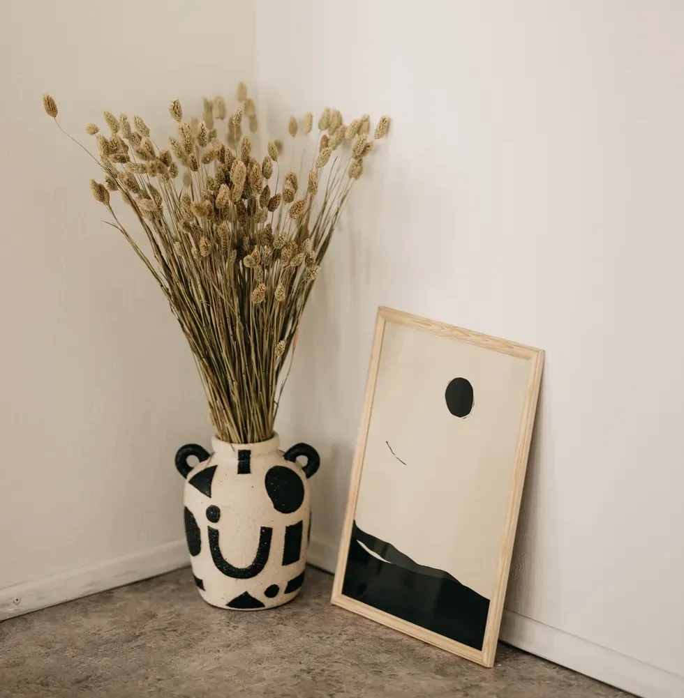 An abstract, black and white painting sits on the ground next to a vase with plants in it.