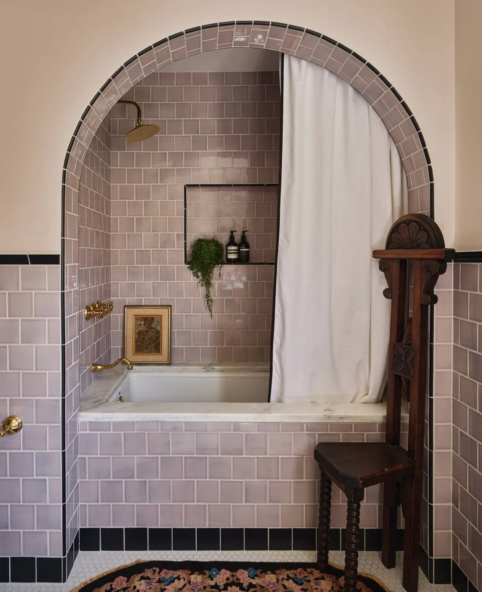 Arched lavender-tiled bathroom with tub, gold fixtures, curtain, and antique wooden chair.