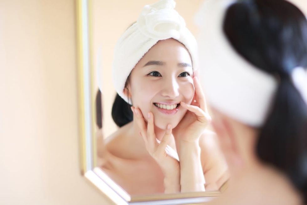 Asian woman touching her face and smiling at her reflection in the mirror during her skincare routine.