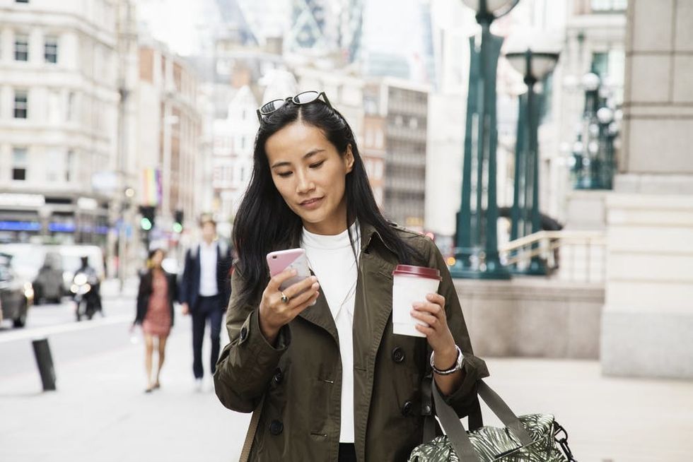 Asian woman walking through city carrying coffee and scrolling on phone.