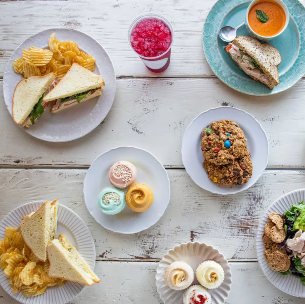Assorted sandwiches, chips, drinks, cookies, cupcakes, and macarons on a white wooden table.