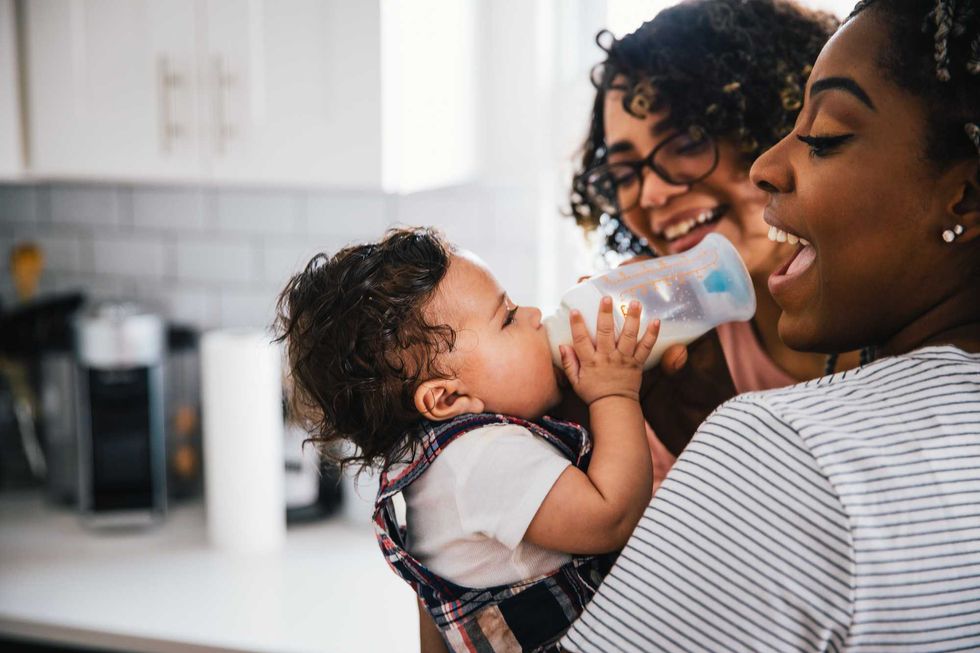 Baby drinking from a bottle, held by two smiling women in a kitchen.