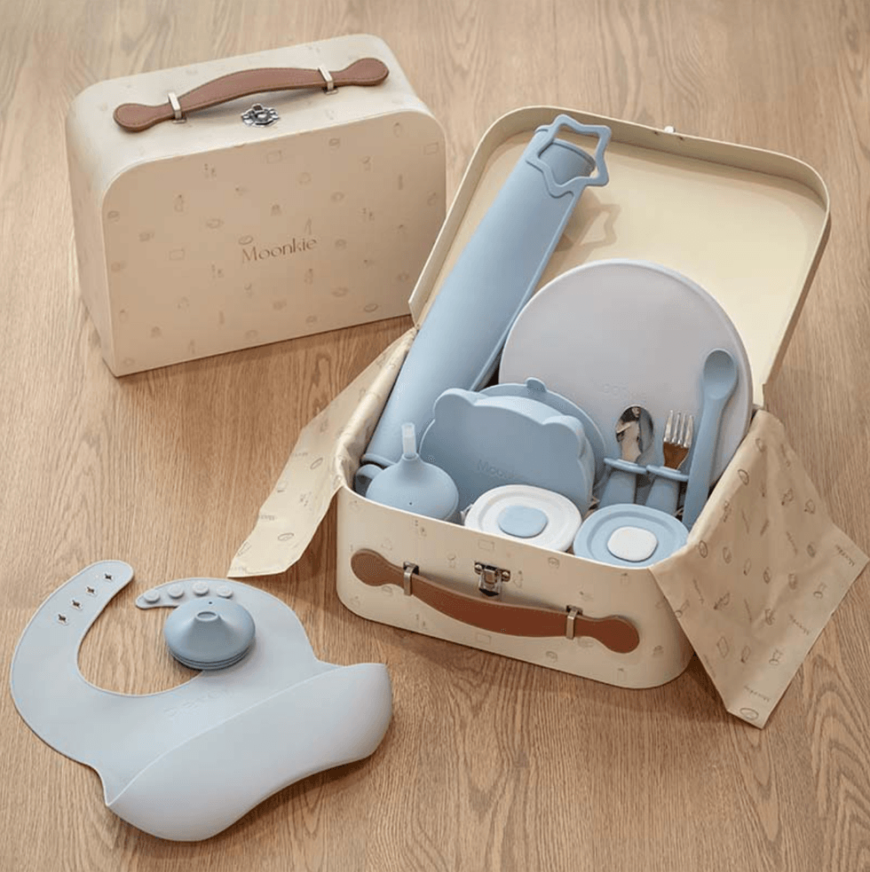 Baby feeding set with utensils and bib in an open case on a wooden floor.
