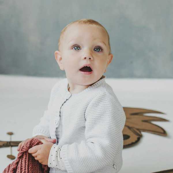 Baby holding a pink blanket, wearing a white sweater, with a surprised expression.
