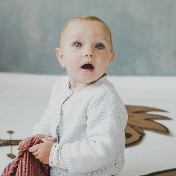 Baby holding a pink blanket, wearing a white sweater, with a surprised expression.