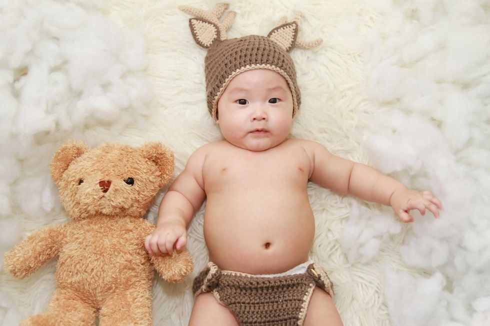 Baby in a deer hat with teddy bear, lying on fluffy white blanket.