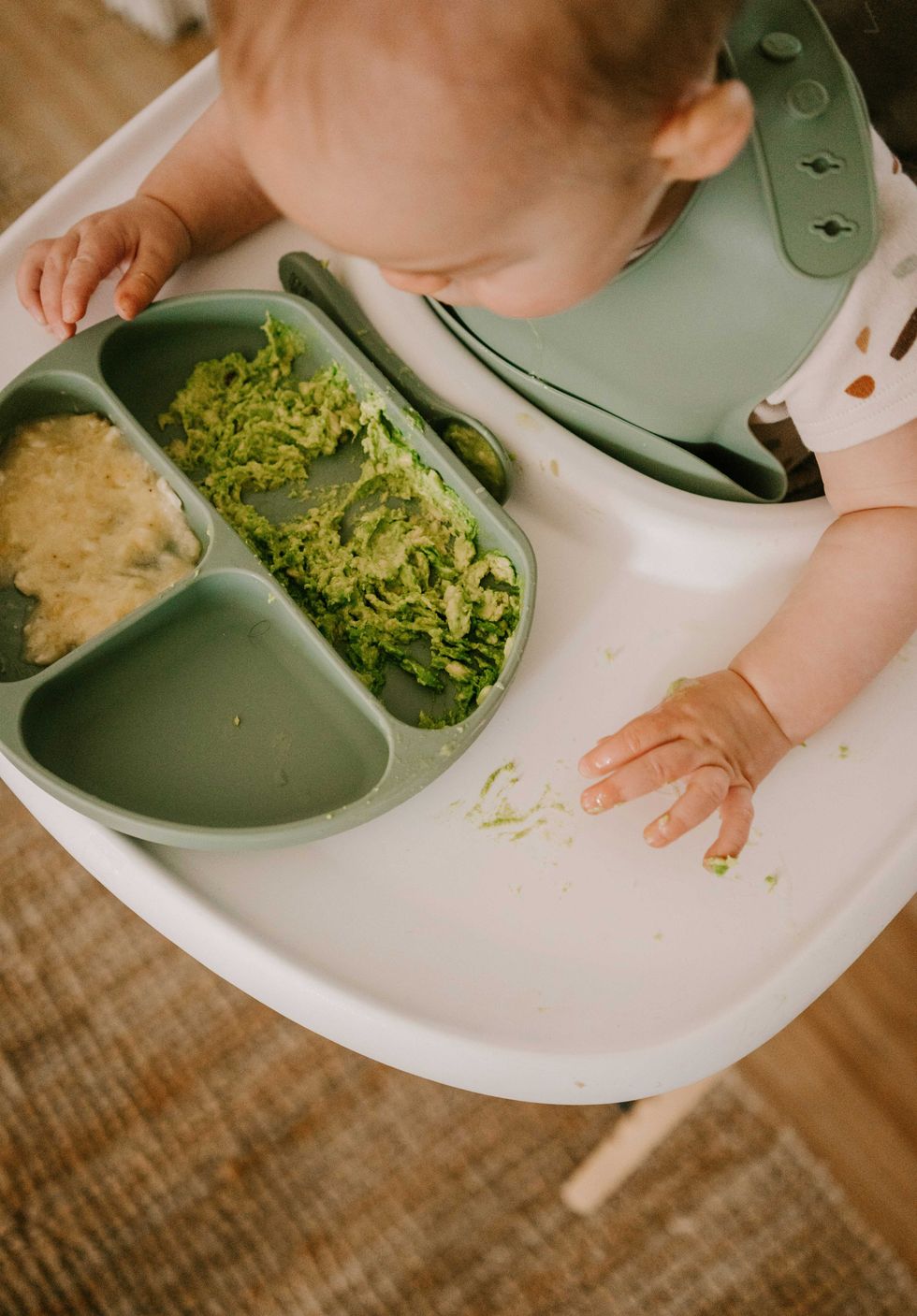 Baby in a highchair with mashed avocado and banana on the tray.