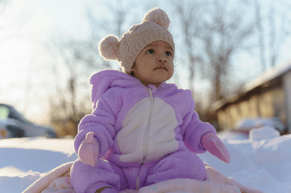 Baby in a purple snowsuit and knit hat sits outside in the snow.