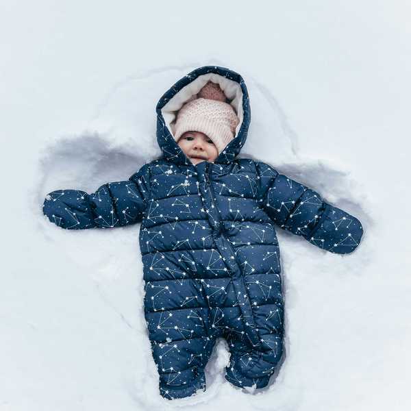 Baby in patterned snowsuit and pink hat lying in snow, making a snow angel.