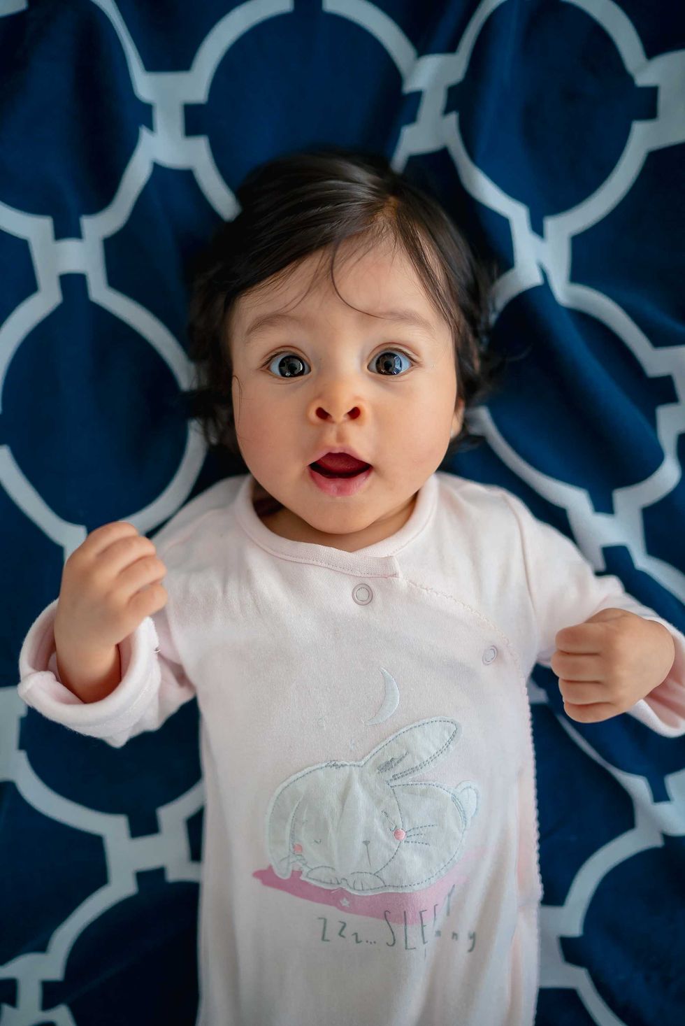 Baby with surprised expression, lying on a blue geometric patterned blanket.