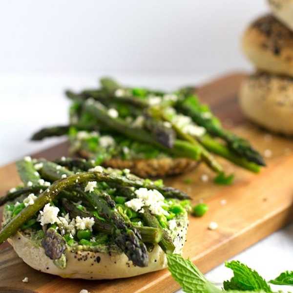 Bagel topped with asparagus, peas, and crumbled cheese on a wooden board.
