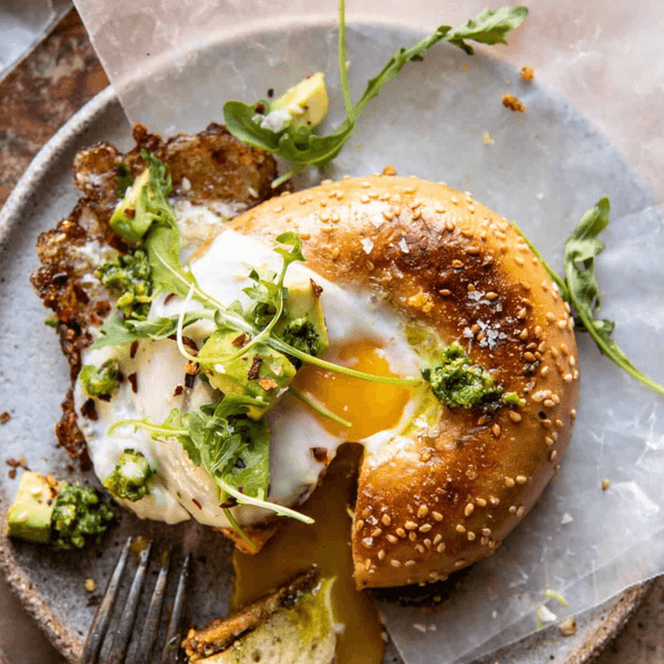 Bagel with fried egg, avocado, arugula, pesto, and a runny yolk on a plate.
