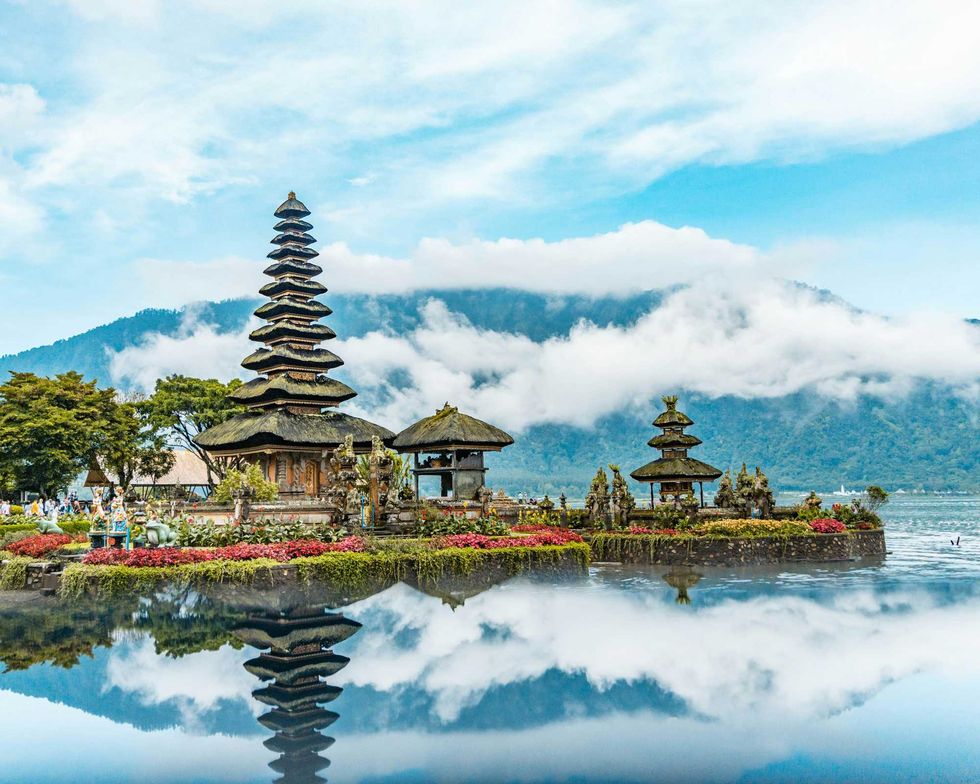Balinese temple with tiered roofs by a lake, reflected in calm water, surrounded by mountains.