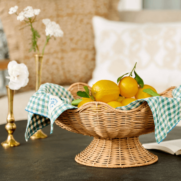 Basket of lemons with blue cloth on table, with flowers and pillows in background.