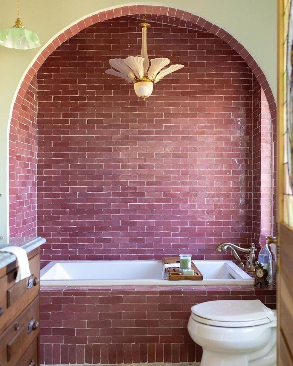 Bathroom with red brick walls, white bathtub, ornate chandelier, and a toilet.