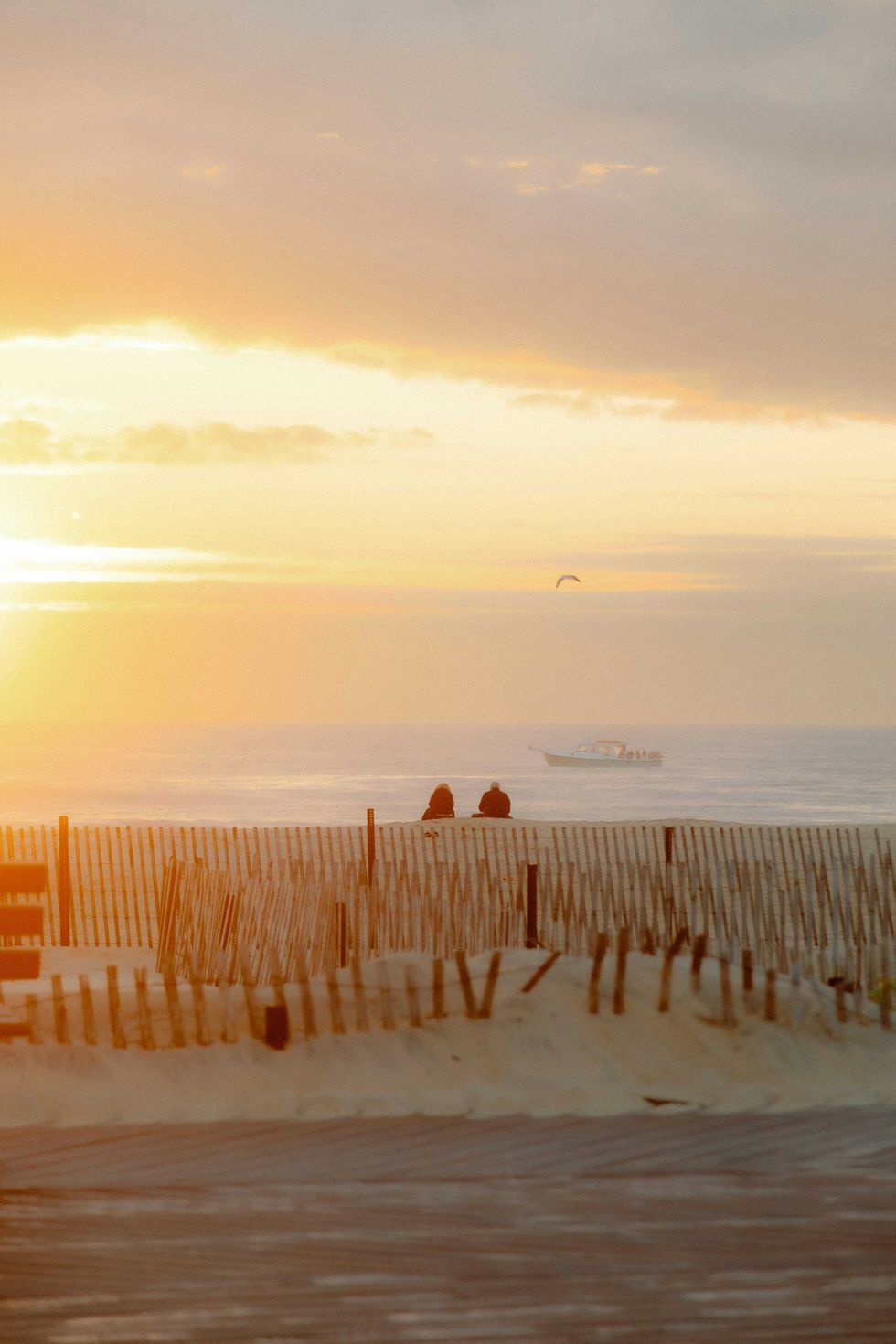 Beach sunrise with two people, a boat, and sand fences in the foreground.
