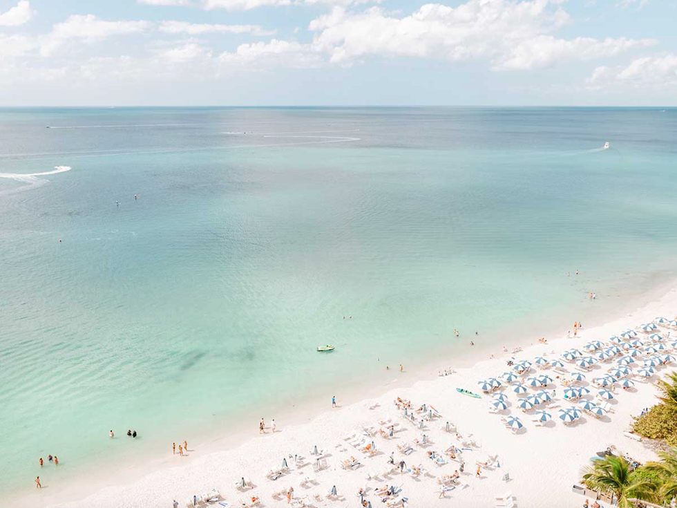 Beach with blue umbrellas, white sand, and people in calm turquoise water.