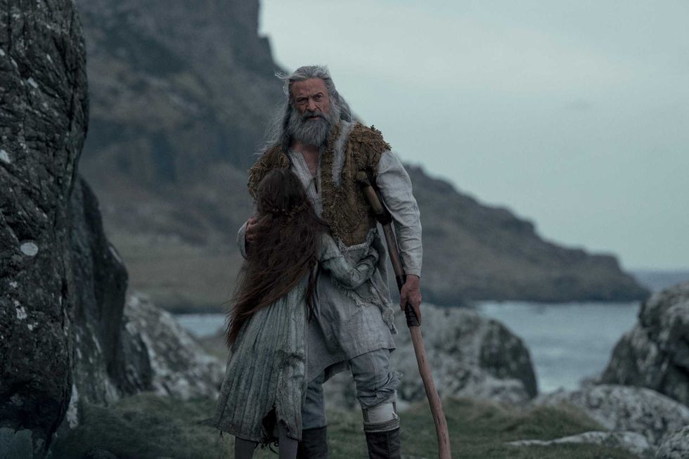 Bearded man in rustic clothing holds a staff by rocky seaside landscape.