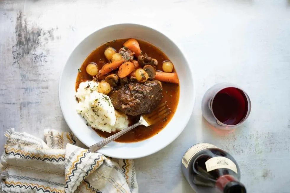 Beef bourguignon sits in a bowl with a fork, next to a glass of wine.